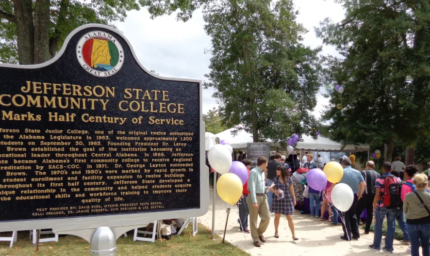 A large crowd gathers near a Jefferson State Community College sign during an outdoor event. People hold balloons in white, yellow, and purple, and tents are set up in the background under tall trees.
