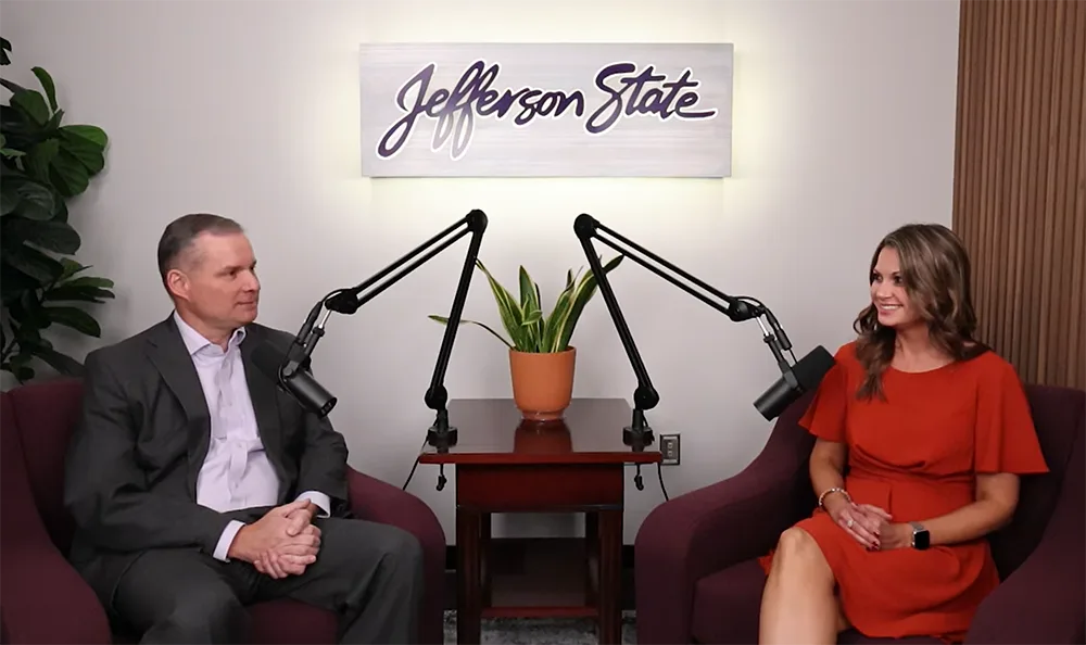 A man in a suit and a woman in an orange dress sit in armchairs with microphones between them, having a conversation. A sign reading Jefferson State hangs on the wall behind them.