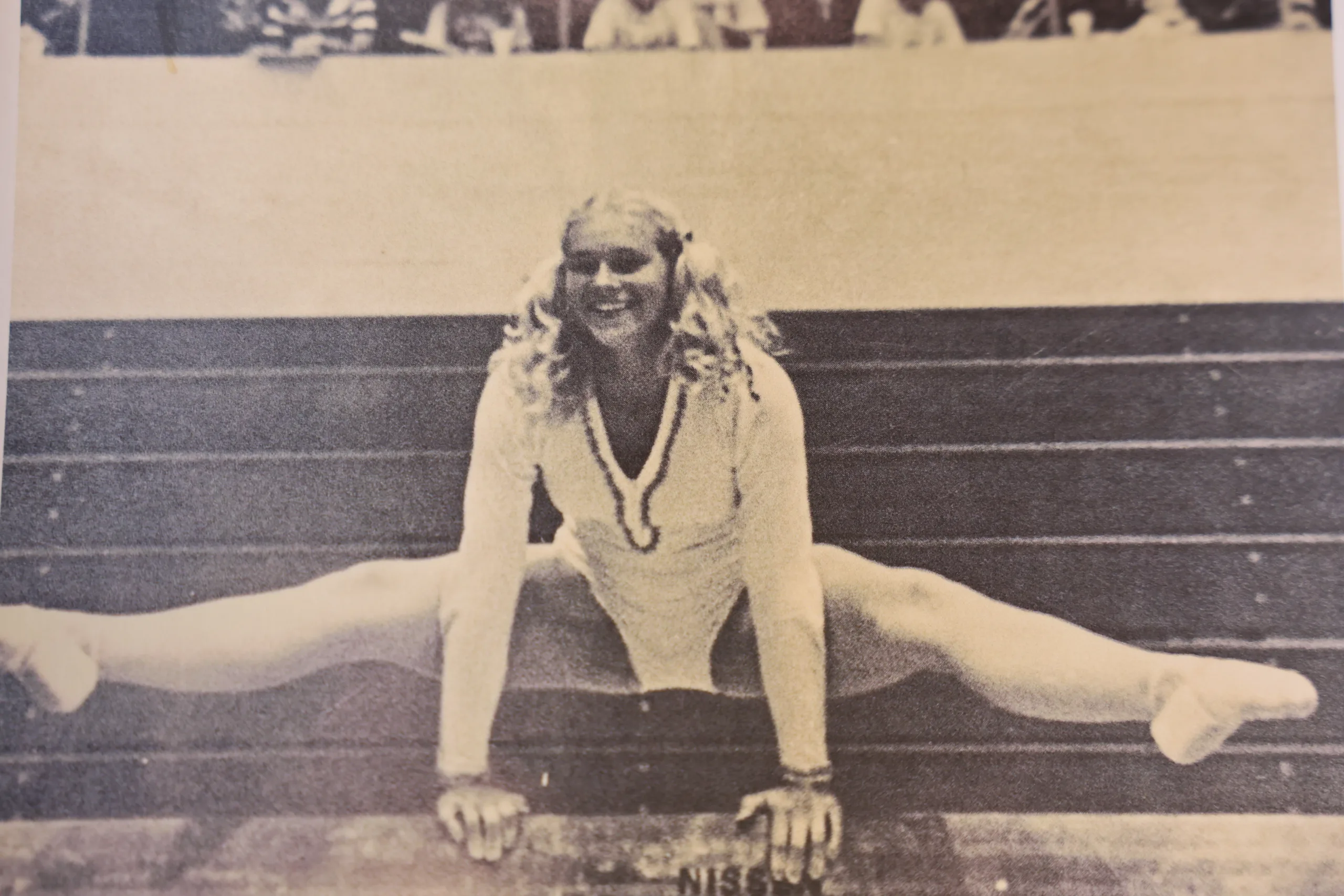 A gymnast in a leotard and socks performs a split on parallel bars, smiling at the camera. The image is in black and white and appears to be vintage.