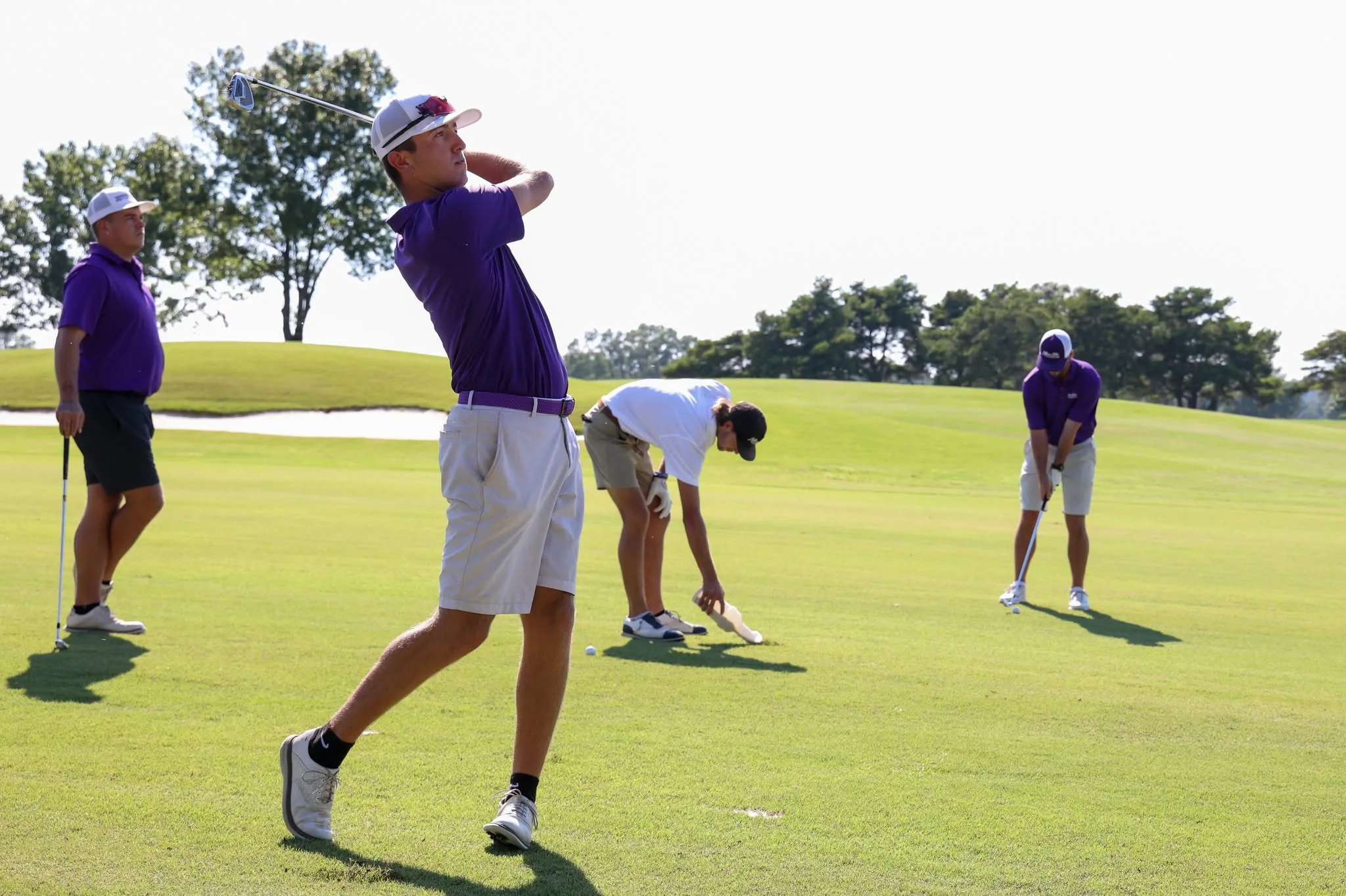 Four golfers are on a green golf course. One golfer in the foreground swings a club, while the others in the background are preparing to putt or set up their shots. All wear shorts and caps under a clear sky.