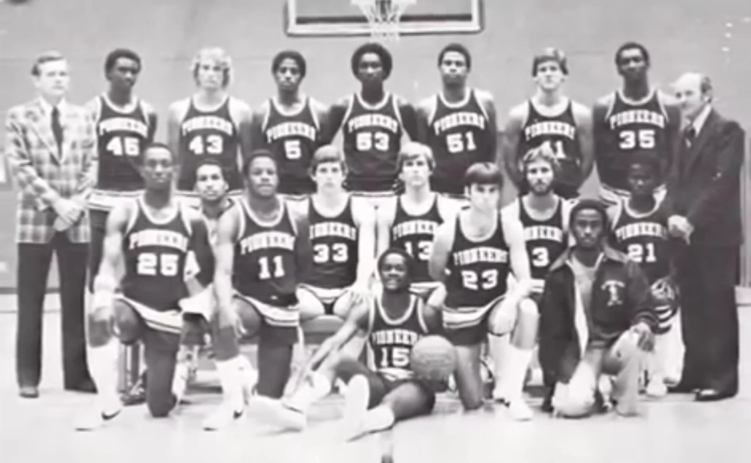 Black-and-white photo of a men’s basketball team posing in uniforms labeled “Pioneers.” The group is arranged in three rows with two coaches in suits standing at each end. A basketball is on the floor in front.