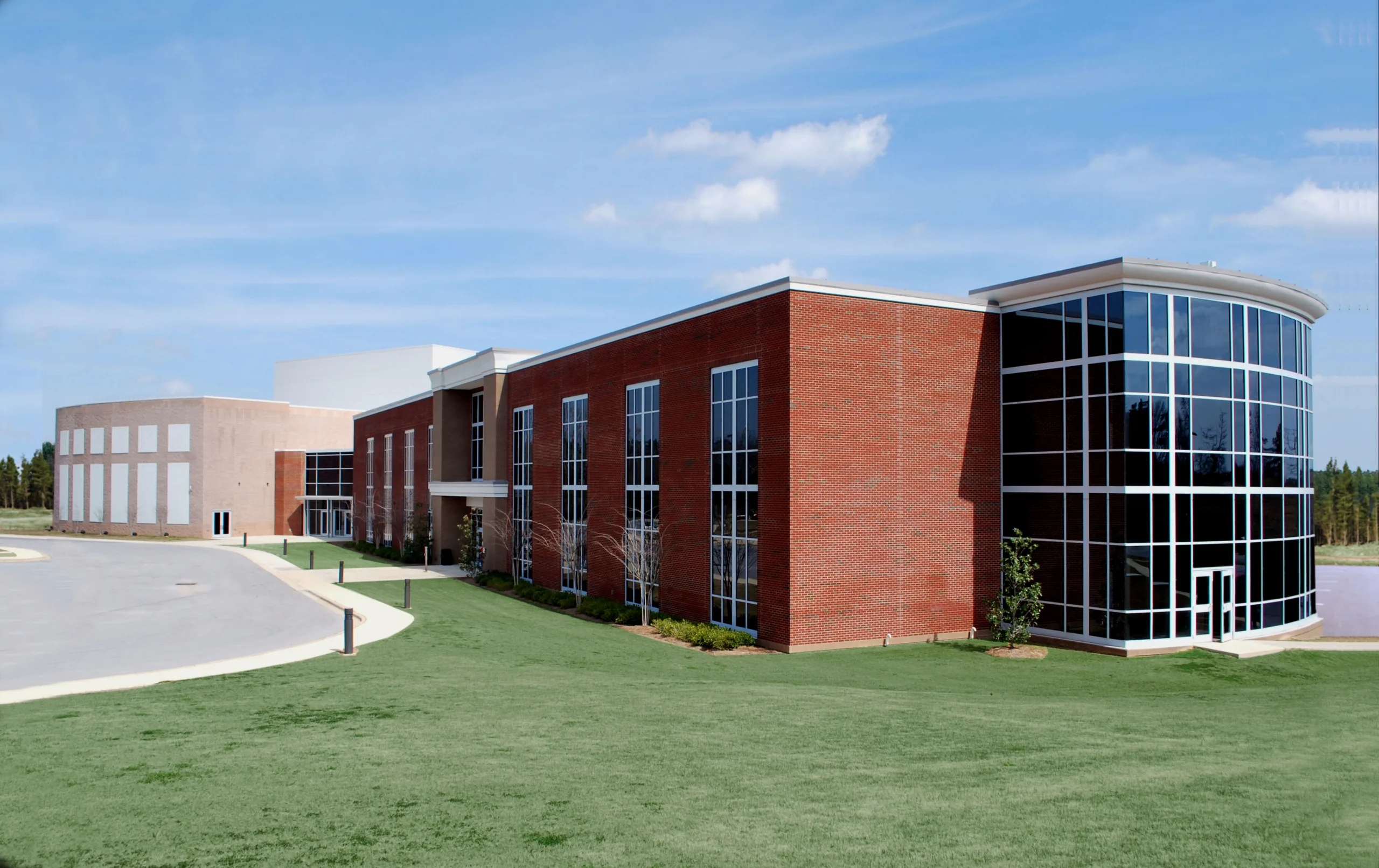 A modern building with red brick walls, large glass windows, and a curved glass entrance, surrounded by a well-manicured lawn under a blue sky.