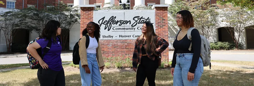 Four students with backpacks stand and talk in front of a Jefferson State Community College sign at the Shelby-Hoover campus, with a brick building and trees in the background.