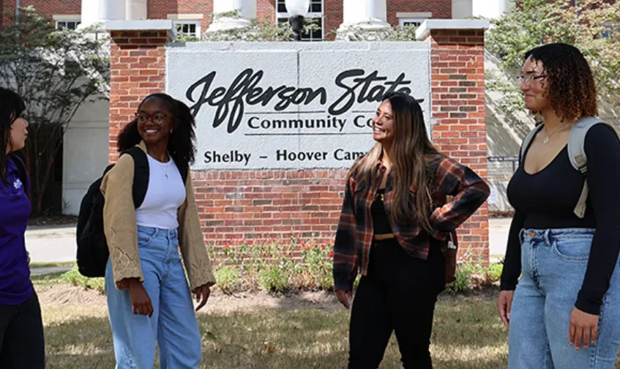Four students with backpacks stand and talk in front of a Jefferson State Community College sign at the Shelby-Hoover campus, with a brick building and trees in the background.