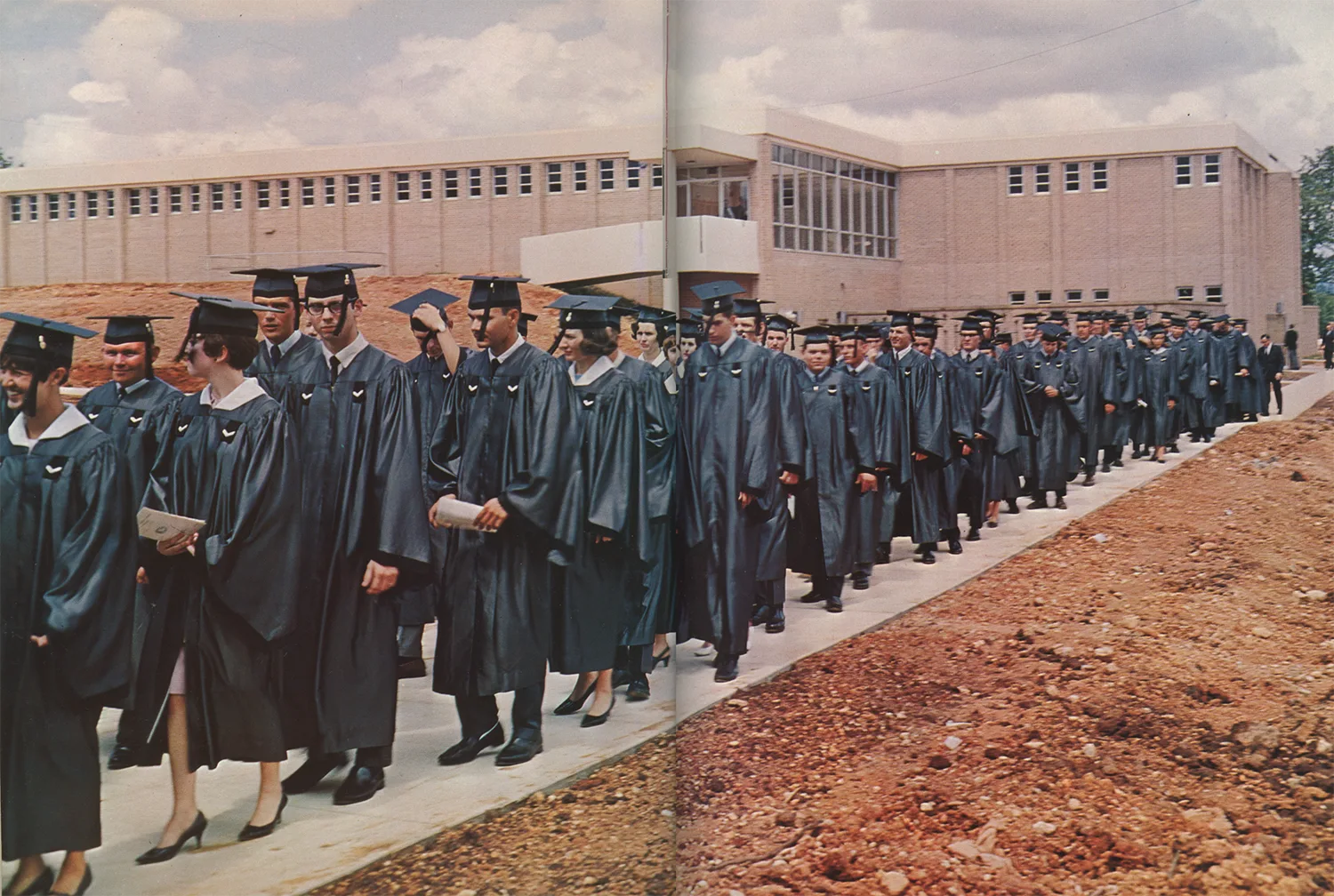 A large group of graduates in black caps and gowns walks in a procession outside a brick school building under a partly cloudy sky. Some hold diplomas, and a dirt area is visible beside the walkway.