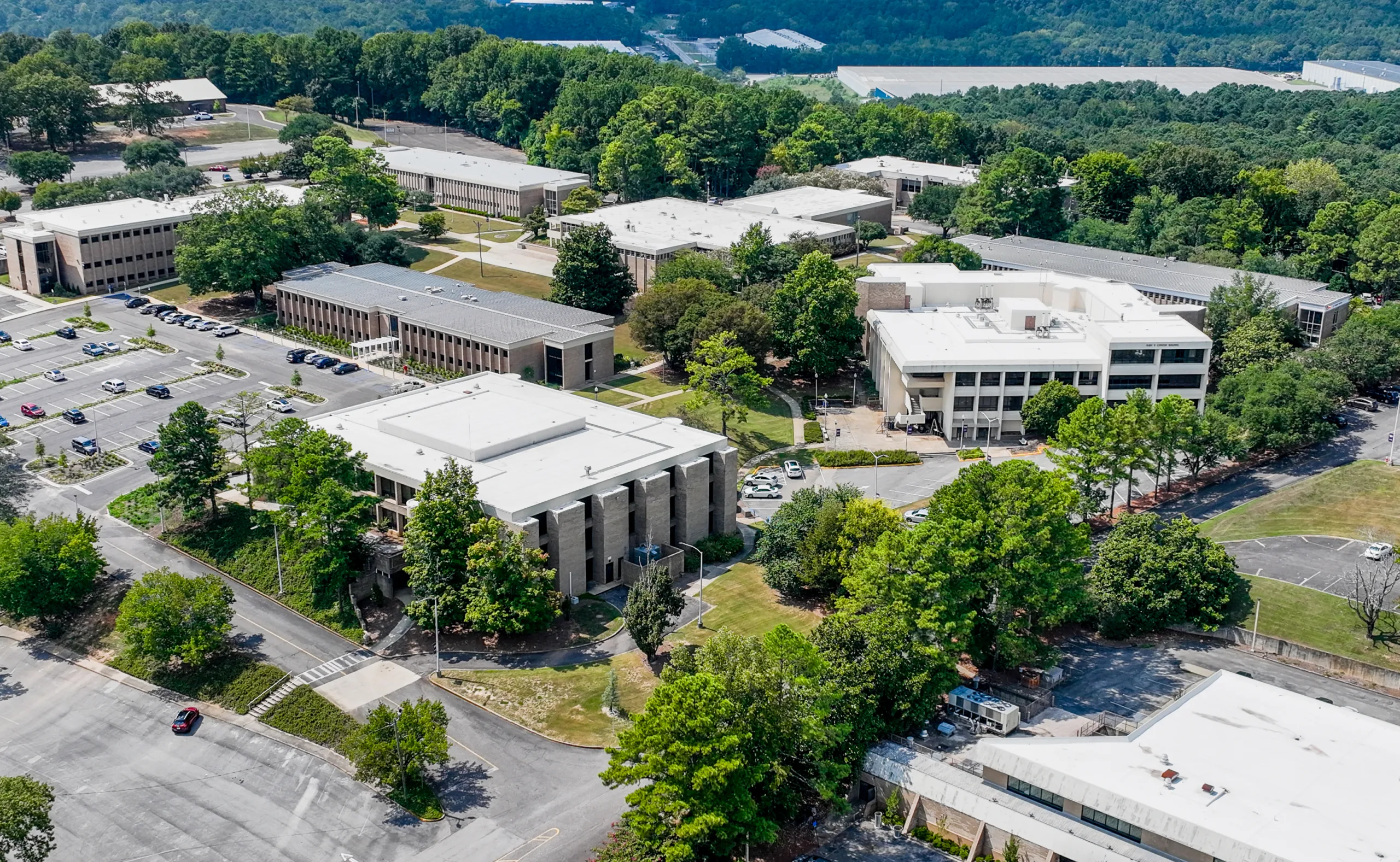 Aerial view of a college campus with several academic buildings, parking lots, and surrounding trees. The area appears quiet, with few cars in the lots and lush greenery throughout the campus.