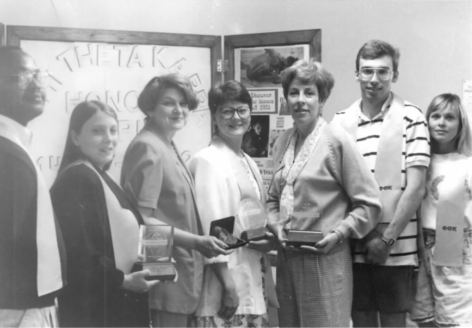 A group of seven people stands indoors, some holding awards. They are smiling and facing the camera. Behind them, a display board is visible with text, including Theta Kappa and Honor.