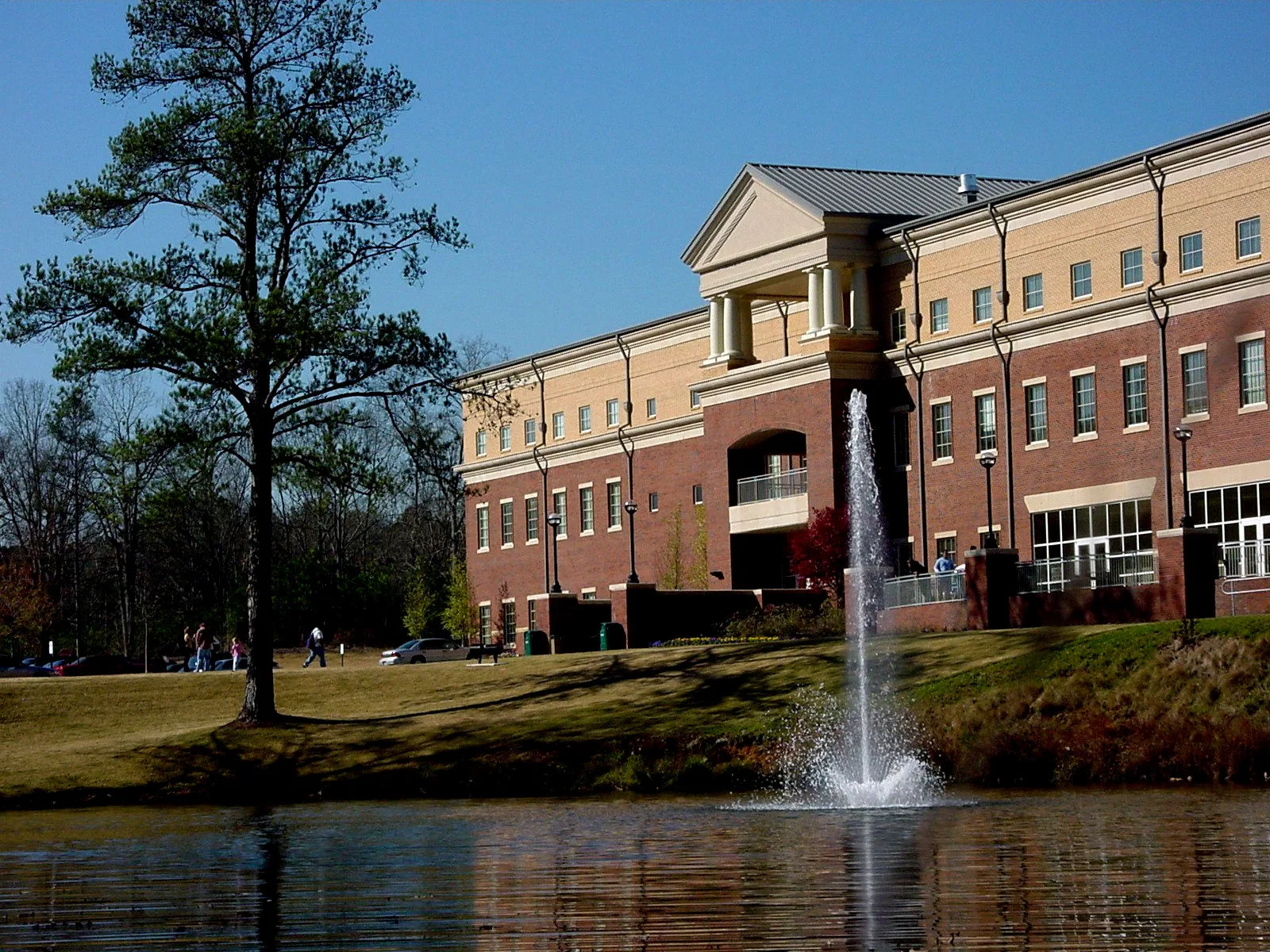 A large brick and tan building with columns stands behind a pond with a water fountain, surrounded by grass, trees, and people walking on a sunny day.