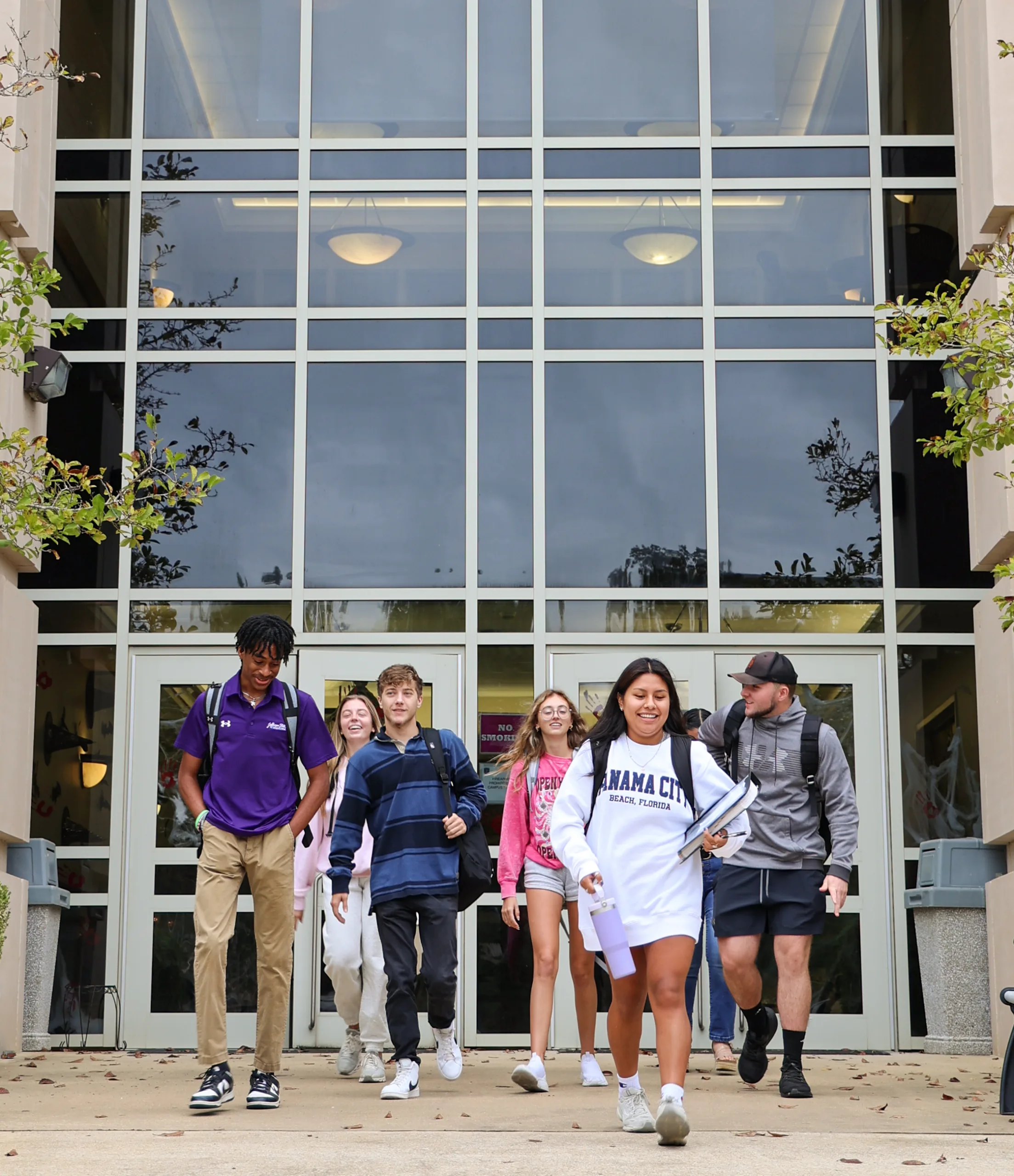 A group of six students, carrying backpacks and wearing casual clothes, walk together out of a modern building with large glass windows. They appear to be chatting and smiling.