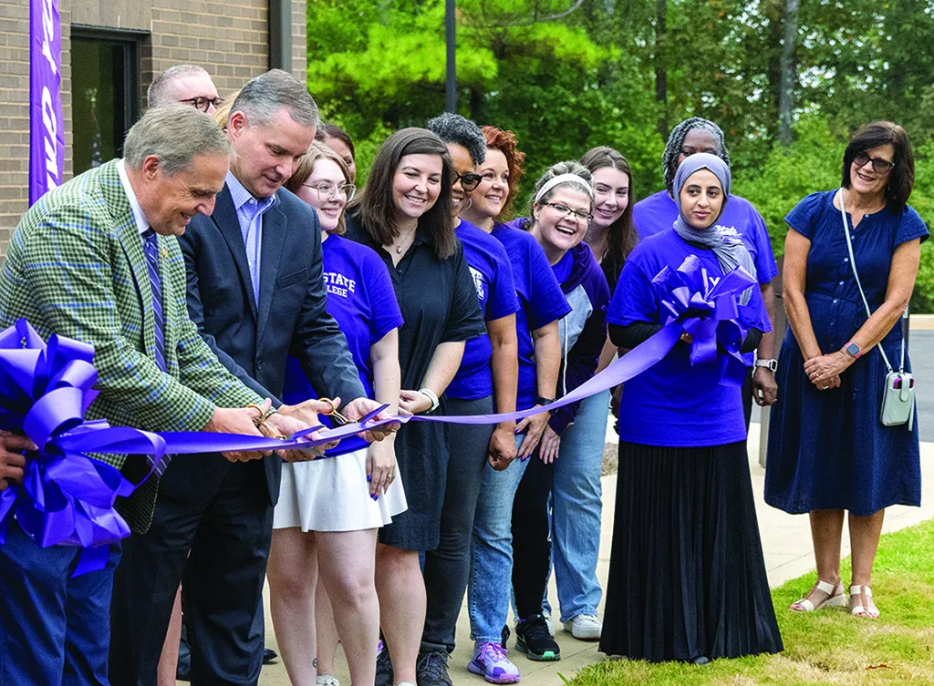 A group of people, some in purple shirts, smile during a ribbon-cutting ceremony outdoors. Two men at the front hold and cut a large purple ribbon with oversized scissors, while others look on happily.