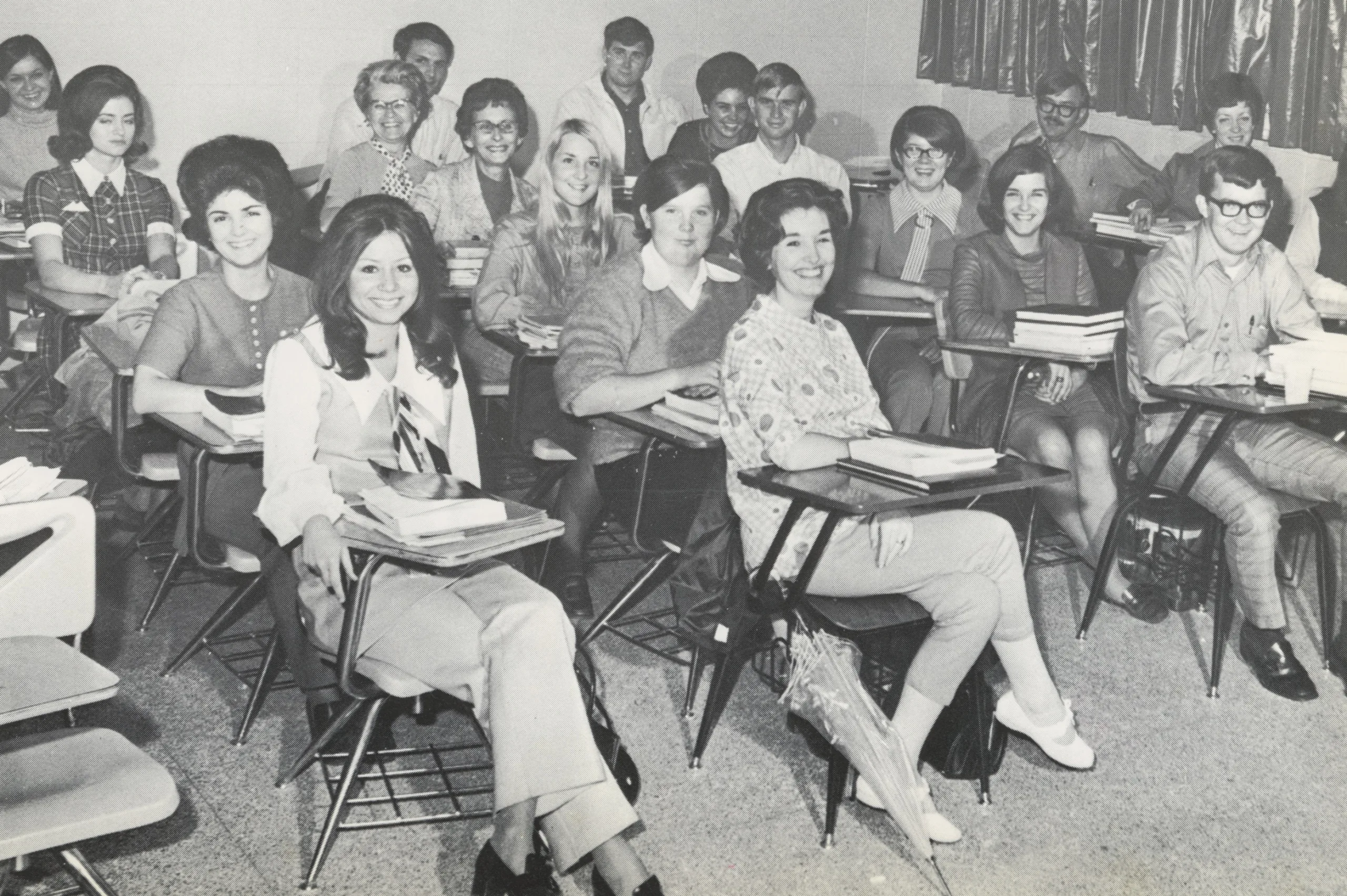 A black and white photo of a classroom with students and a teacher sitting at desks, smiling at the camera. The students appear to be young adults, and stacks of books are visible on their desks.