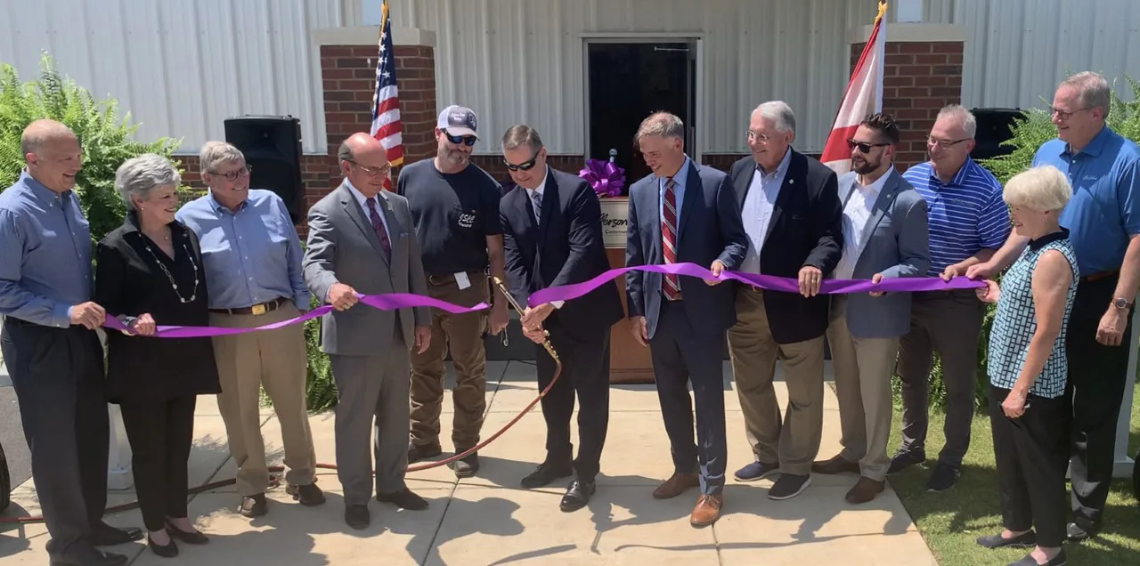 A group of people stands in a line outside a building, holding a long purple ribbon while a man in the center cuts it with large scissors at a ribbon-cutting ceremony. Two flags are visible in the background.