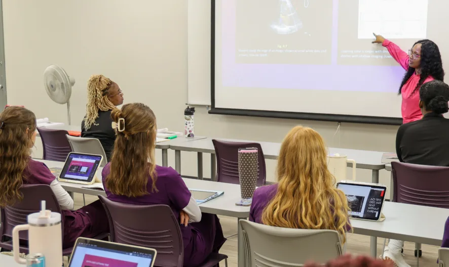 A teacher in pink points at a projected screen while students in purple scrubs sit at desks with laptops, attentively watching in a classroom.