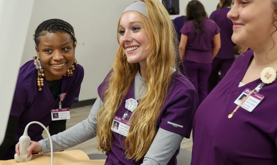Three women in purple scrubs smile and gather around an ultrasound machine in a medical educational setting, with two more people in similar attire visible in the background.