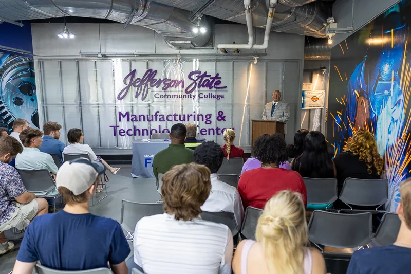 A group of people sit facing a speaker at a podium in the Jefferson State Community College Manufacturing & Technology Center, with banners and industrial decor in the background.